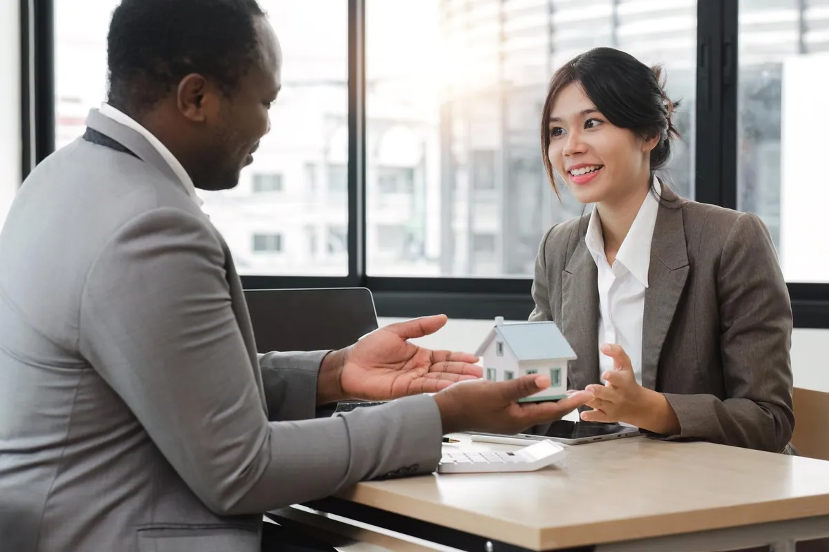 Two professionals in suits talk across a desk while holding a small model house between them